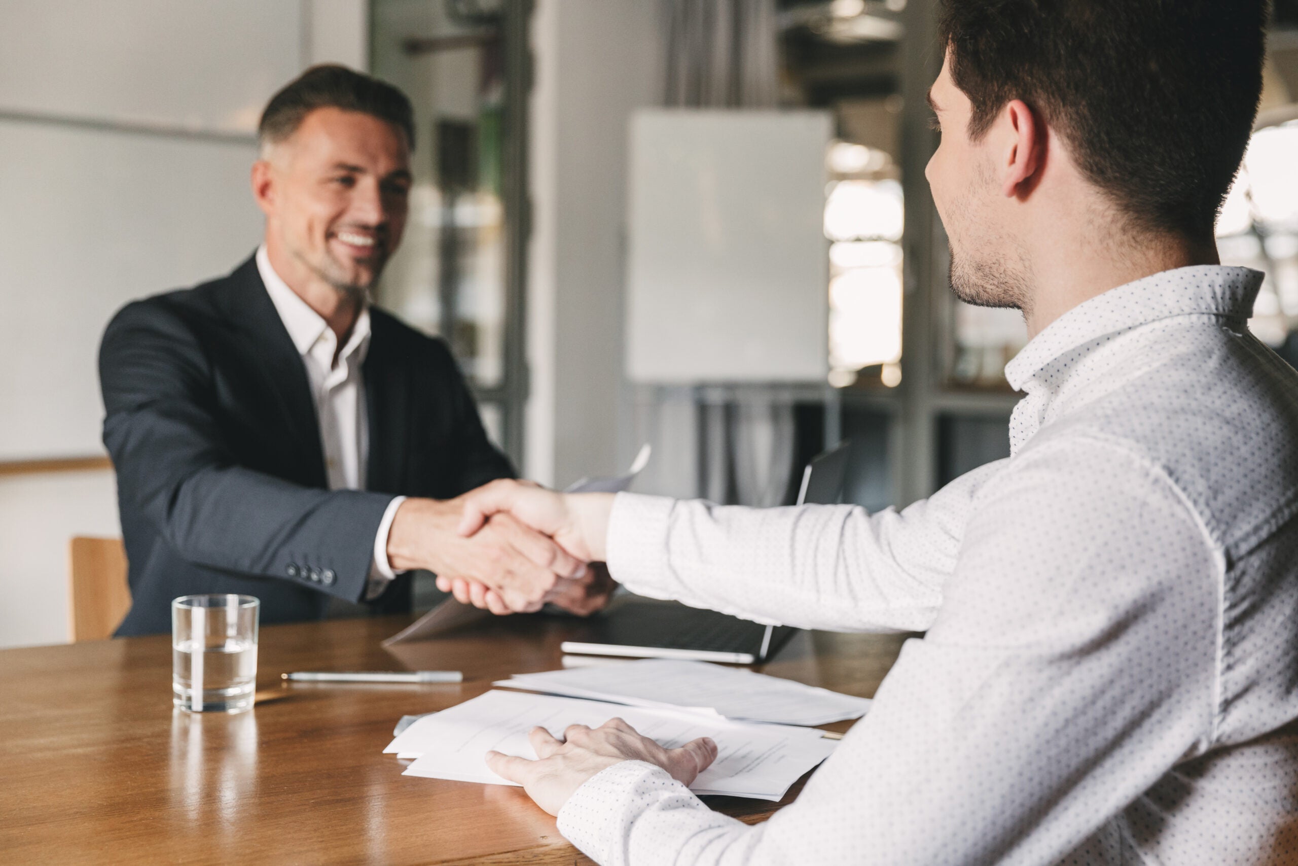 Business, career and placement concept - successful young man smiling and handshaking with european businessman after successful negotiations or interview in office