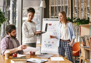 woman standing near whiteboard with a laptop in her hands and looking to her male colleague, that holds a chart with the results. Another colleague looks at it. Concept of success