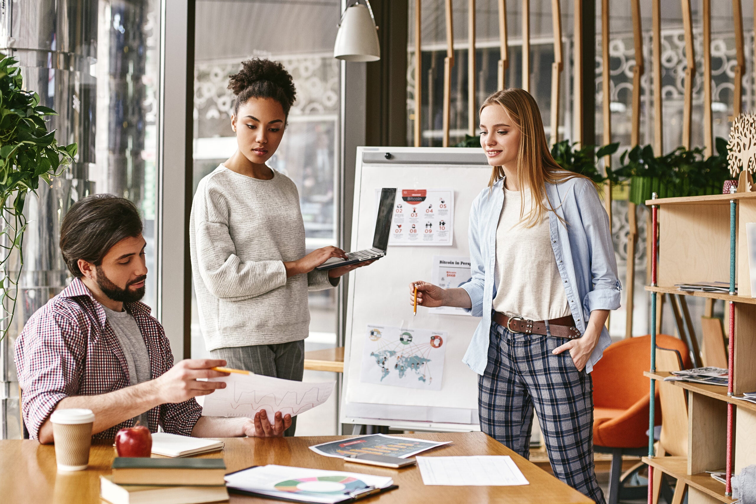 woman standing near whiteboard with a laptop in her hands and looking to her male colleague, that holds a chart with the results. Another colleague looks at it. Concept of success