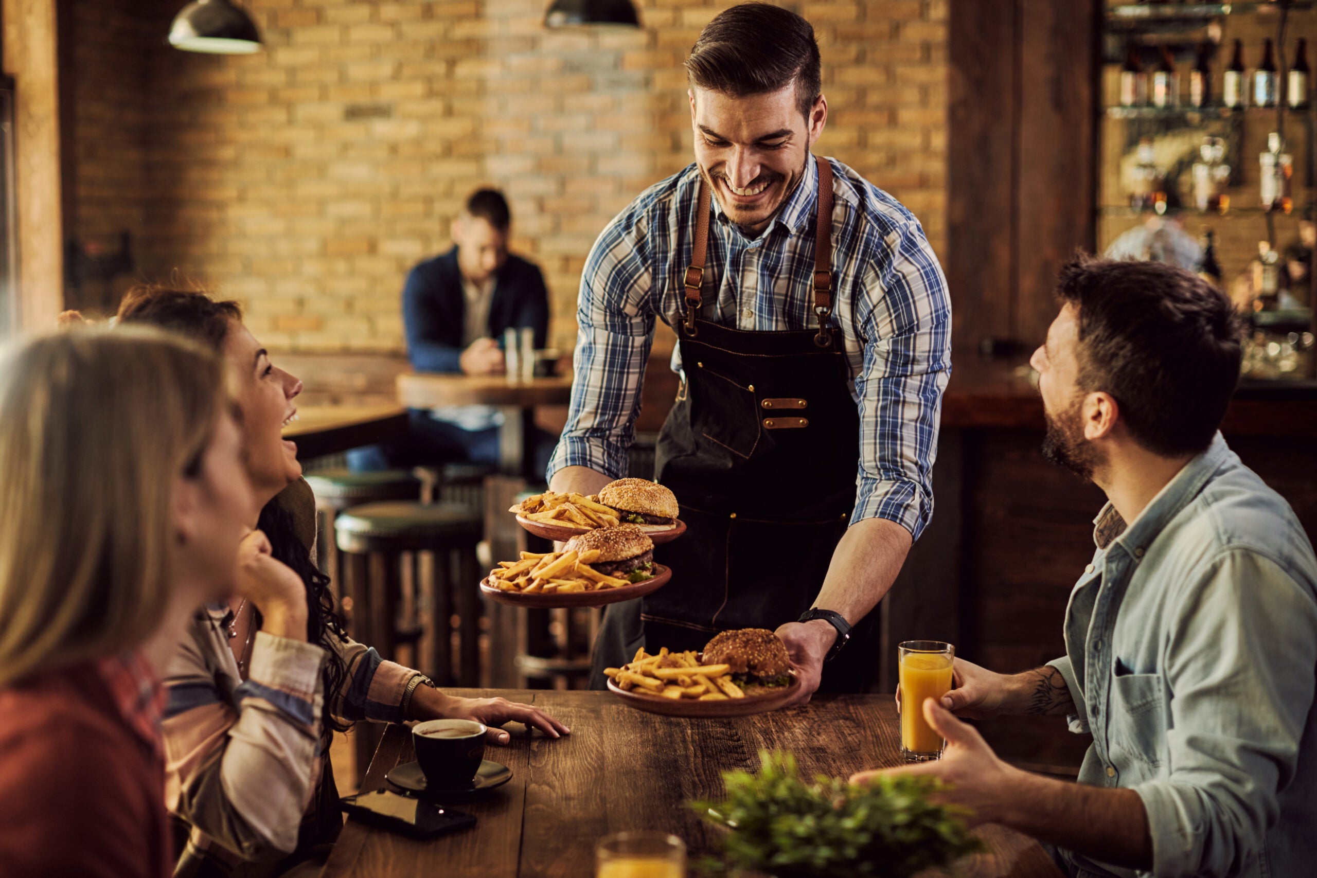 Group of happy friends having fun while waiter is serving them food in a pub. Focus is on waiter.