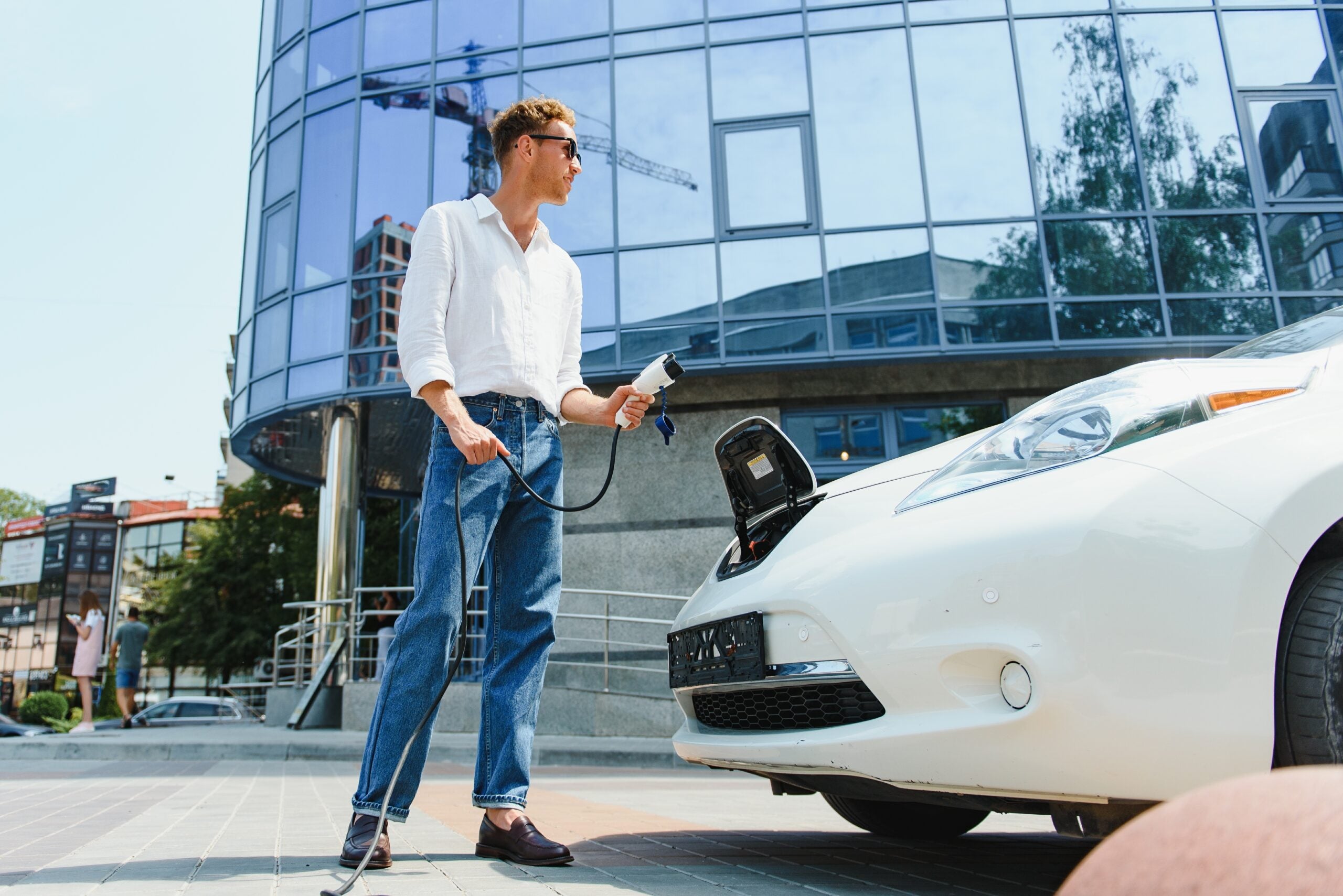 Man Holding Power Charging Cable For Electric Car In Outdoor Car Park. And he s going to connect the car to the charging station in the parking lot near the shopping center