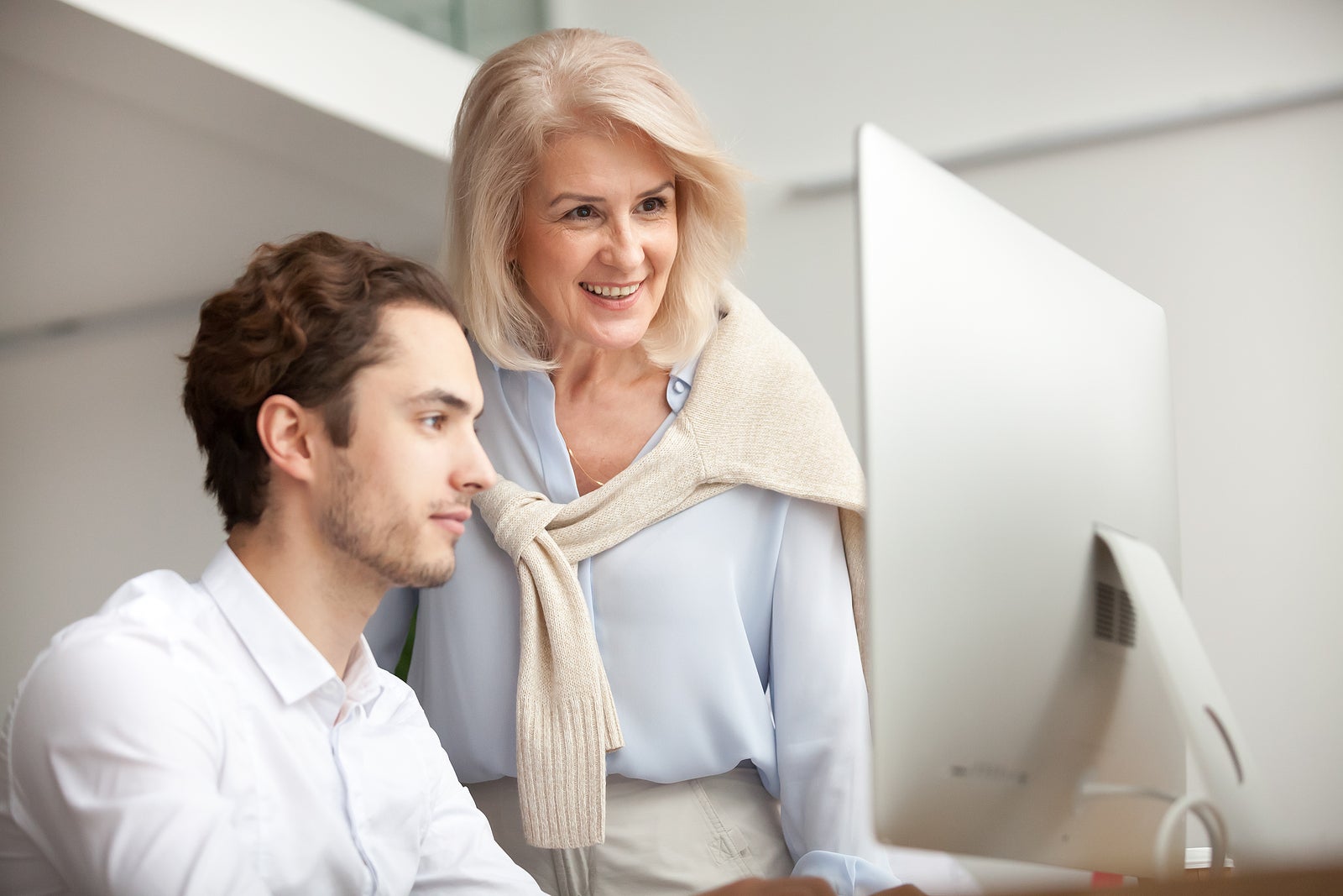 Aged female mentor boss smiling looking at computer screen happy for achievement and good online work result of young male intern, senior woman executive leader supervising teaching helping colleague