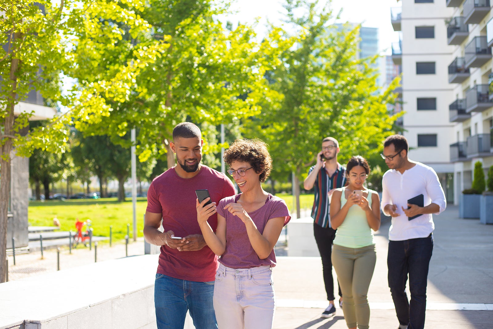 Happy young people using mobile phones while walking through residential area. Men and women talking on cell, using smartphones and showing screen to each other. Wi-Fi outdoors concept